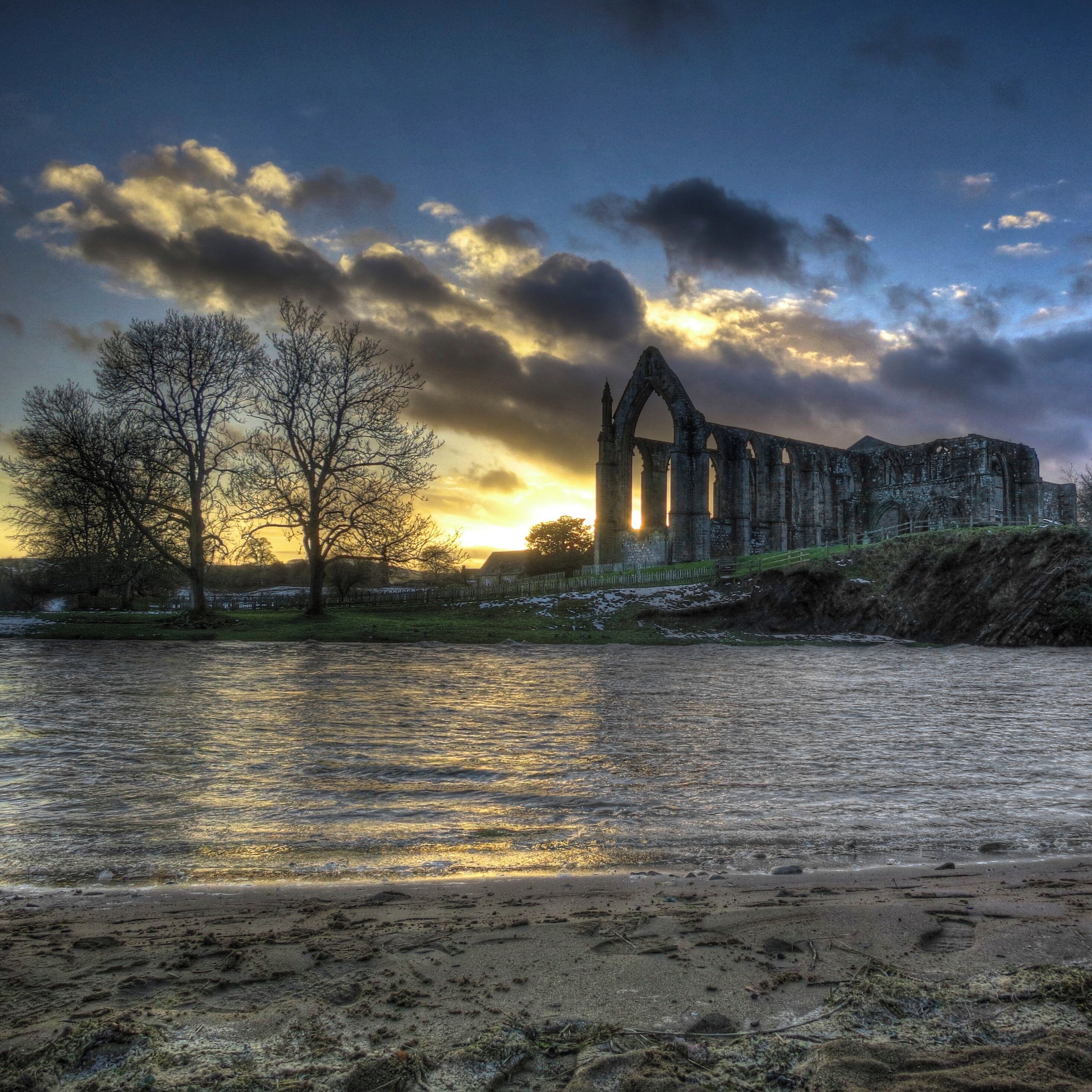 A ruined stone structure by a river with bare trees and a cloudy sky.