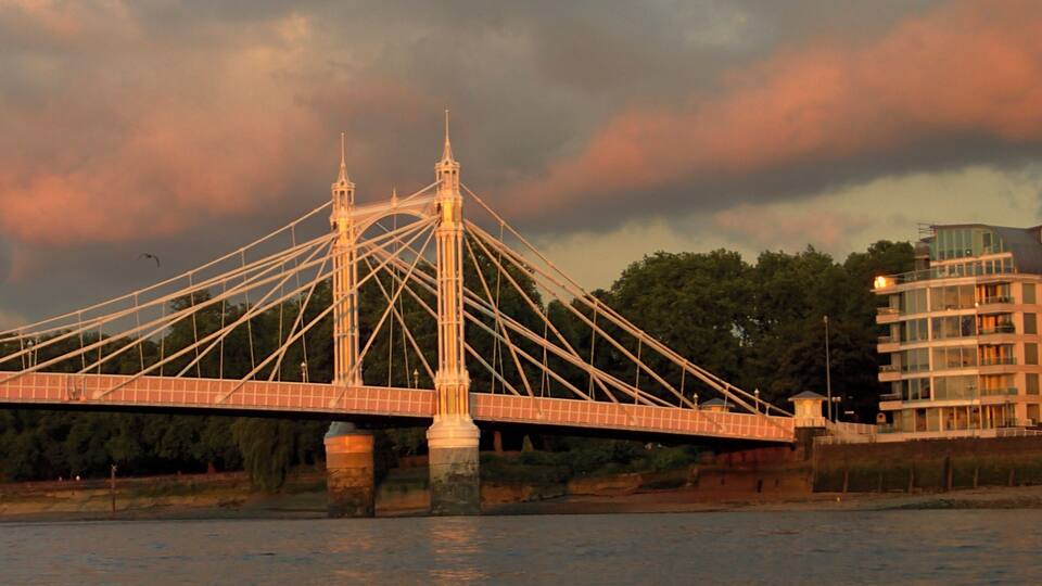 A suspension bridge with two towers, a river below, and a building with balconies in the background.