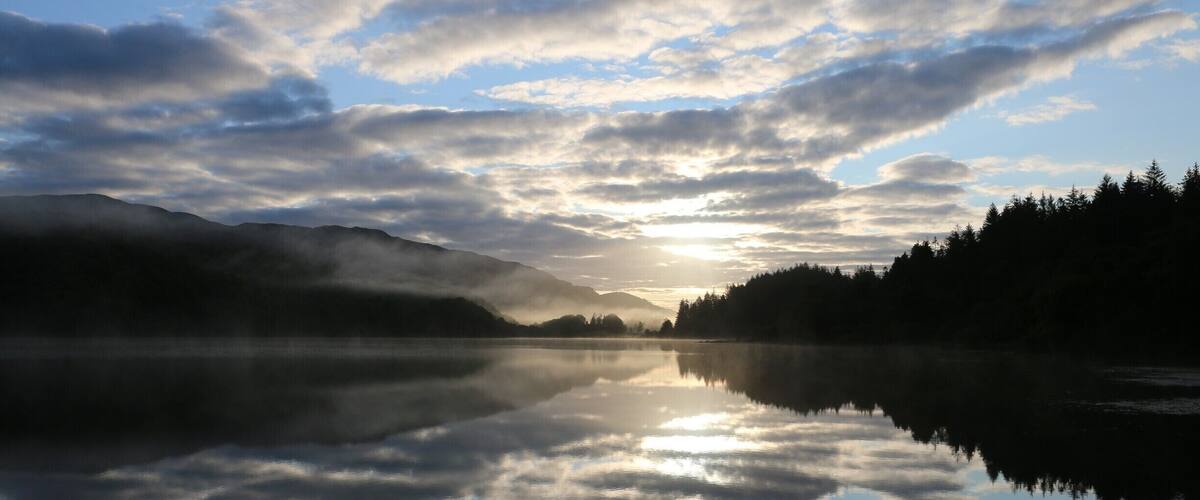 A calm lake reflecting clouds and mountains during sunrise or sunset.