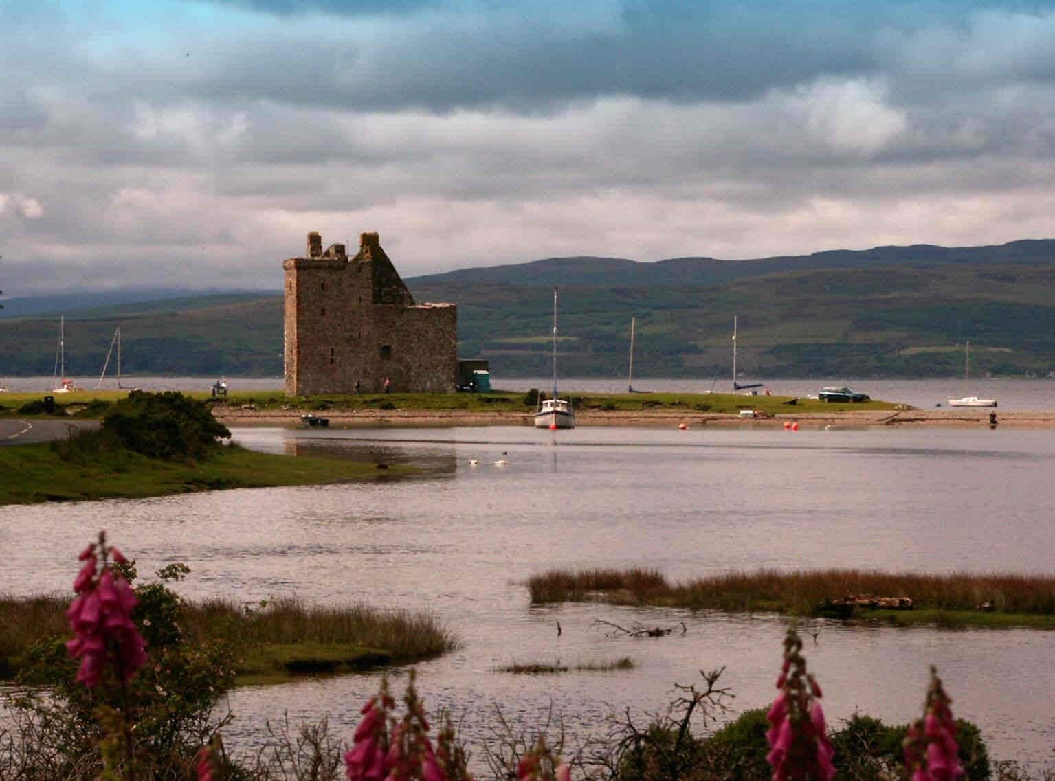 A coastal castle with a tower, surrounded by calm water, boats, and distant hills.