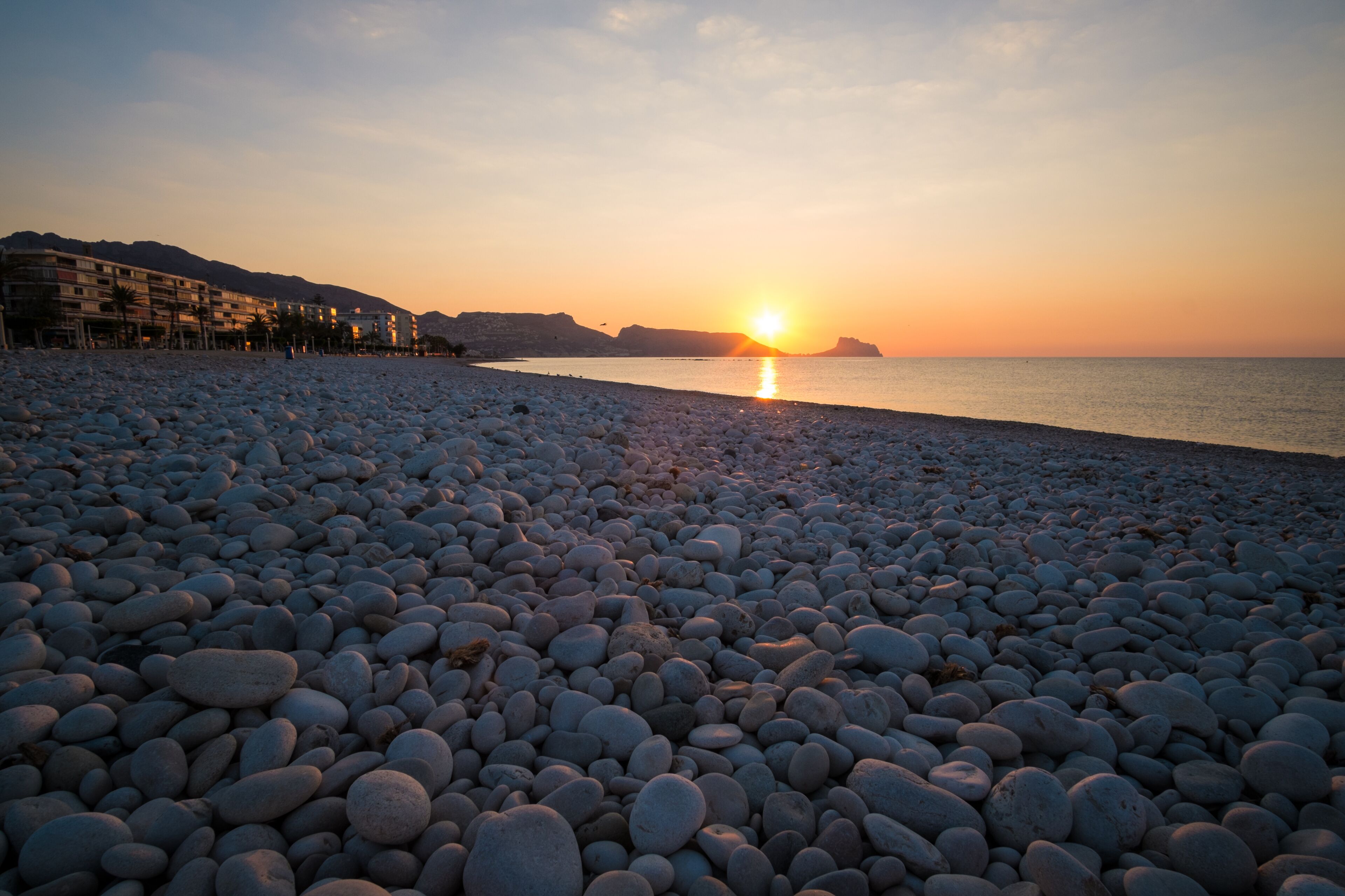A pebble beach with a sunset over the water, buildings in the background, and a distant mountain.