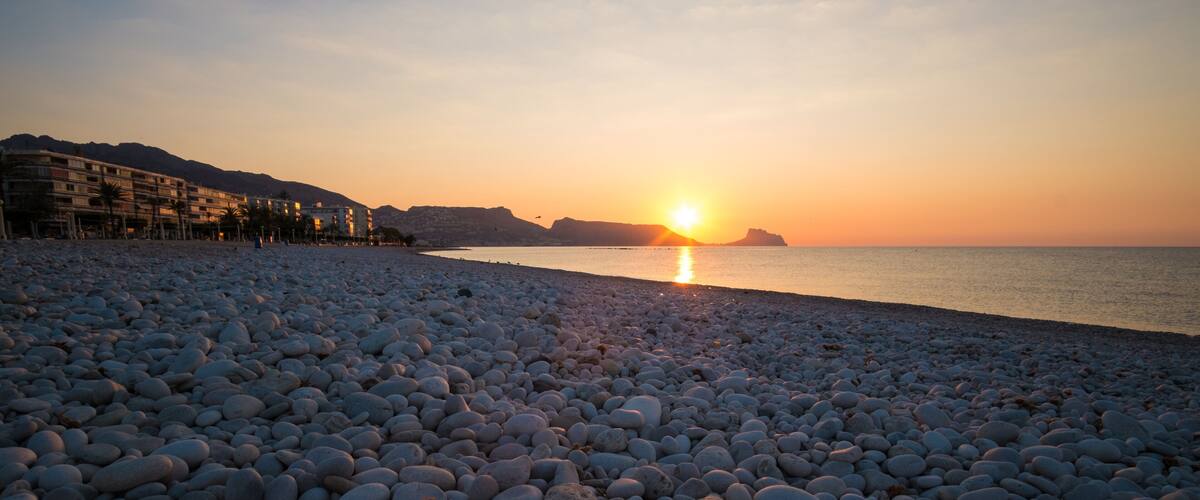 A pebble beach with a sunset over the water, buildings in the background, and a distant mountain.