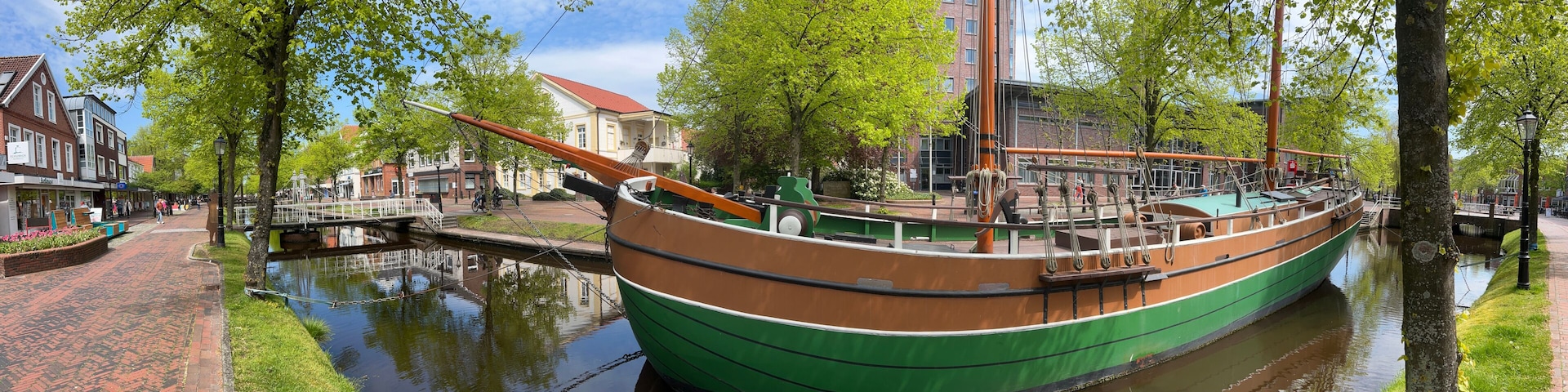 A green and brown boat docked by a canal, surrounded by trees and buildings.