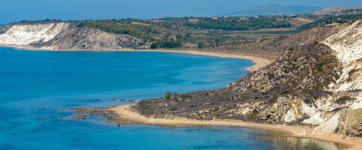 A coastal landscape with a sandy beach, rocky cliffs, and a clear blue sea.