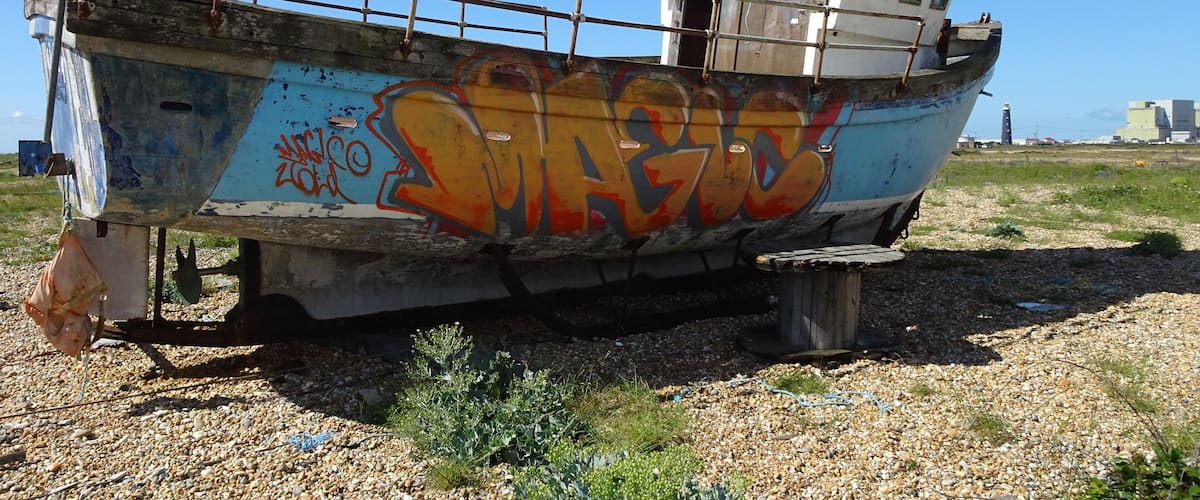 An abandoned boat with graffiti on its side, docked on a gravel beach with a clear sky.