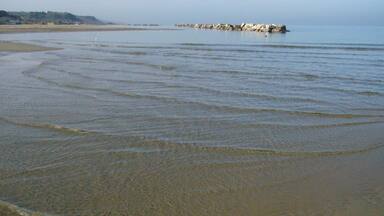 A calm beach with gentle waves, a rocky outcrop in the distance, and a clear sky.