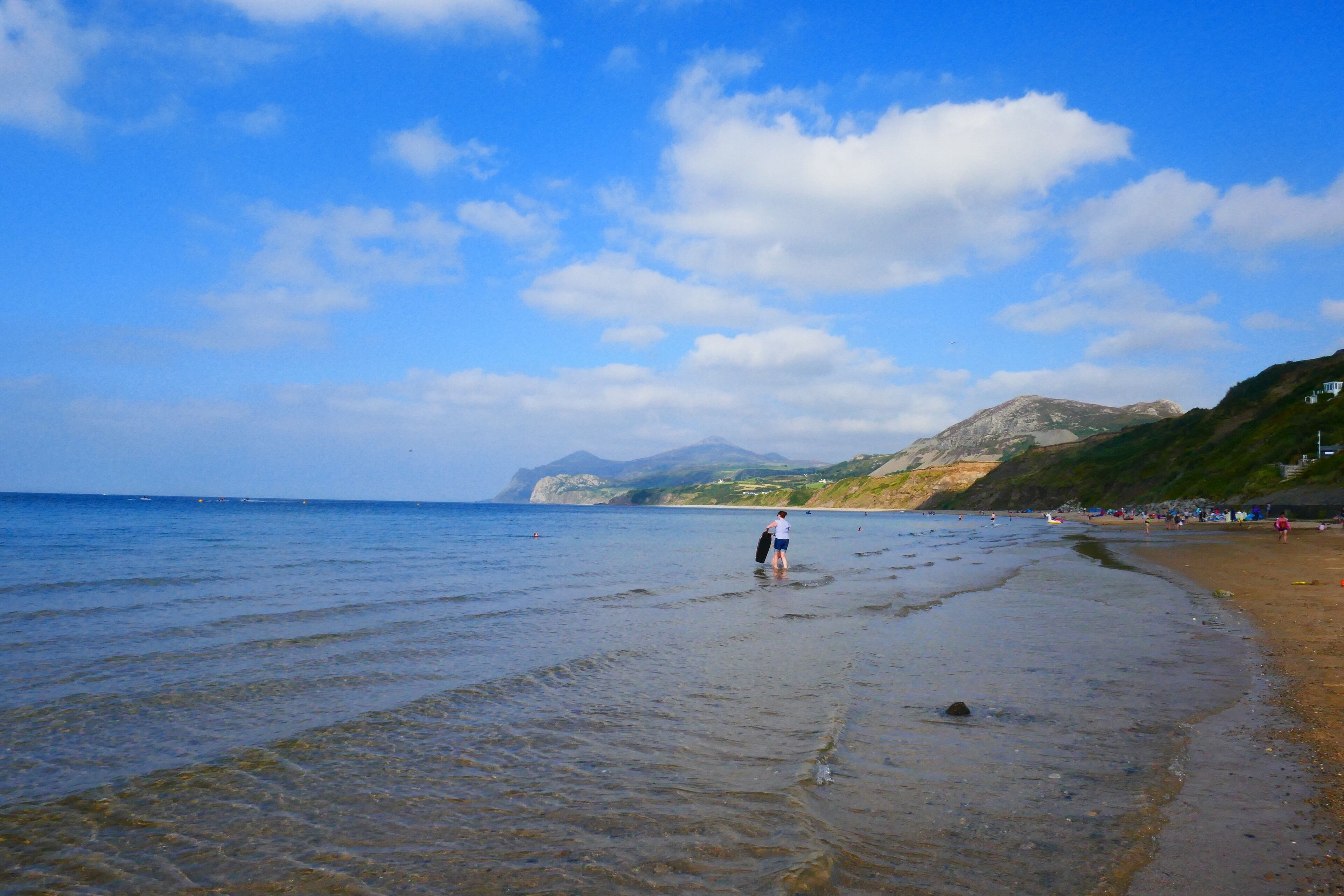 A beach with clear water, a person walking, and distant mountains.