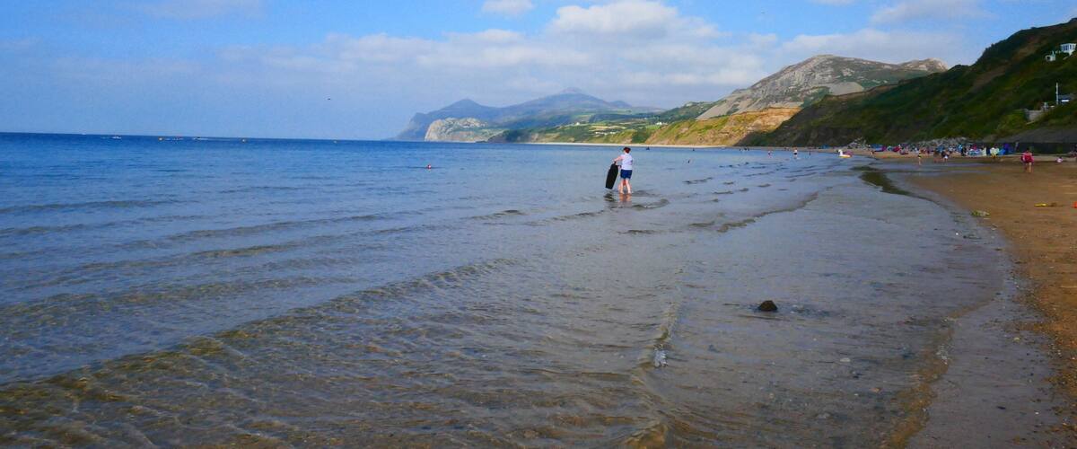 A beach with clear water, a person walking, and distant mountains.