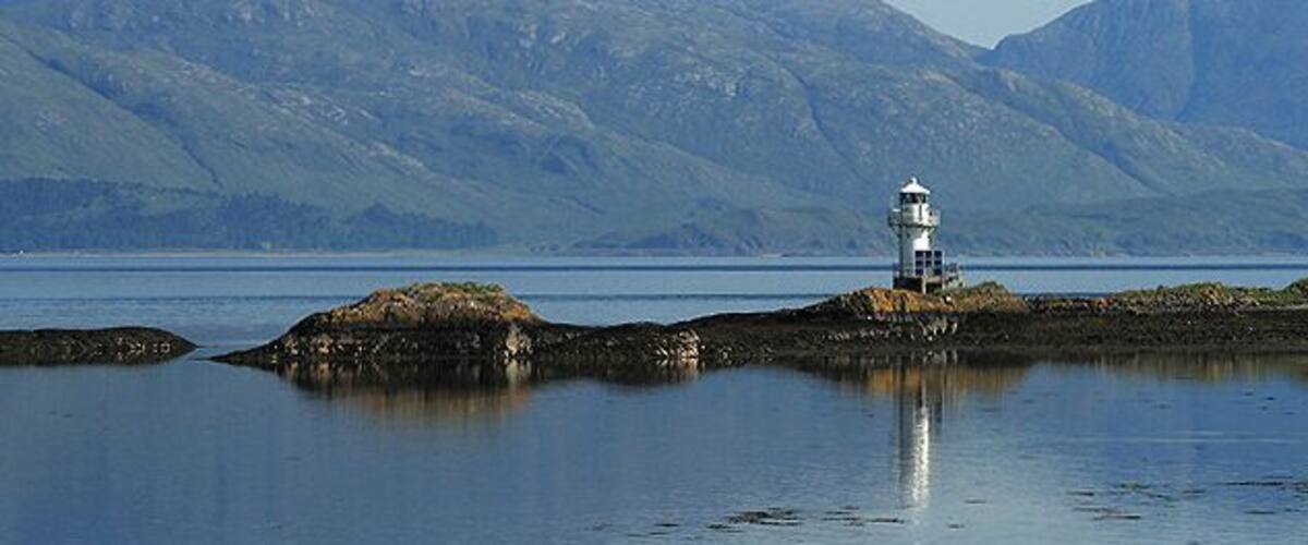 A lighthouse on a rocky outcrop surrounded by calm water and mountains in the background.