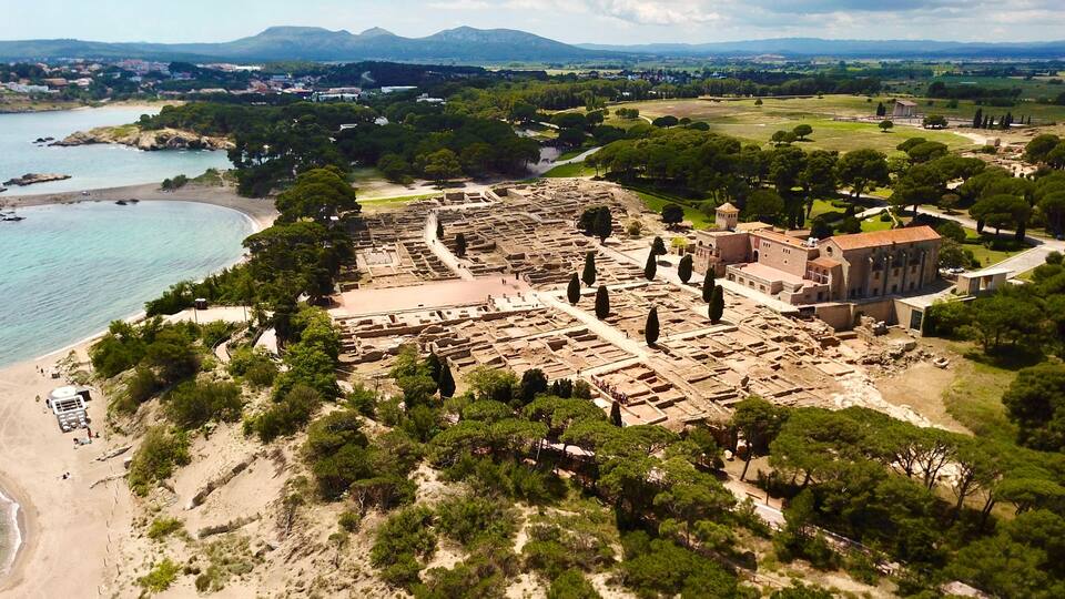 An ancient archaeological site with ruins, a beach, and a mountainous backdrop.