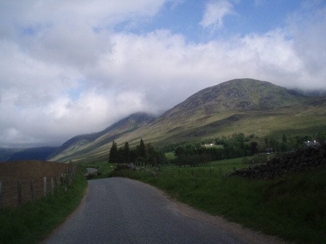 A rural road winding through a hilly landscape with a mountain in the background and a cloudy sky.