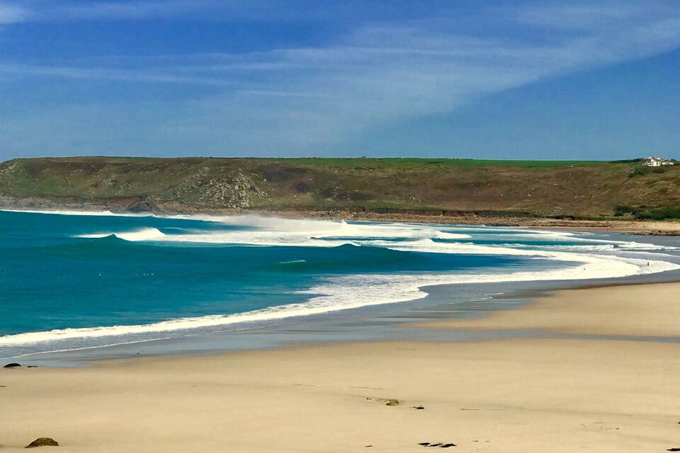 A sandy beach with waves crashing onto the shore and a grassy cliff in the background.