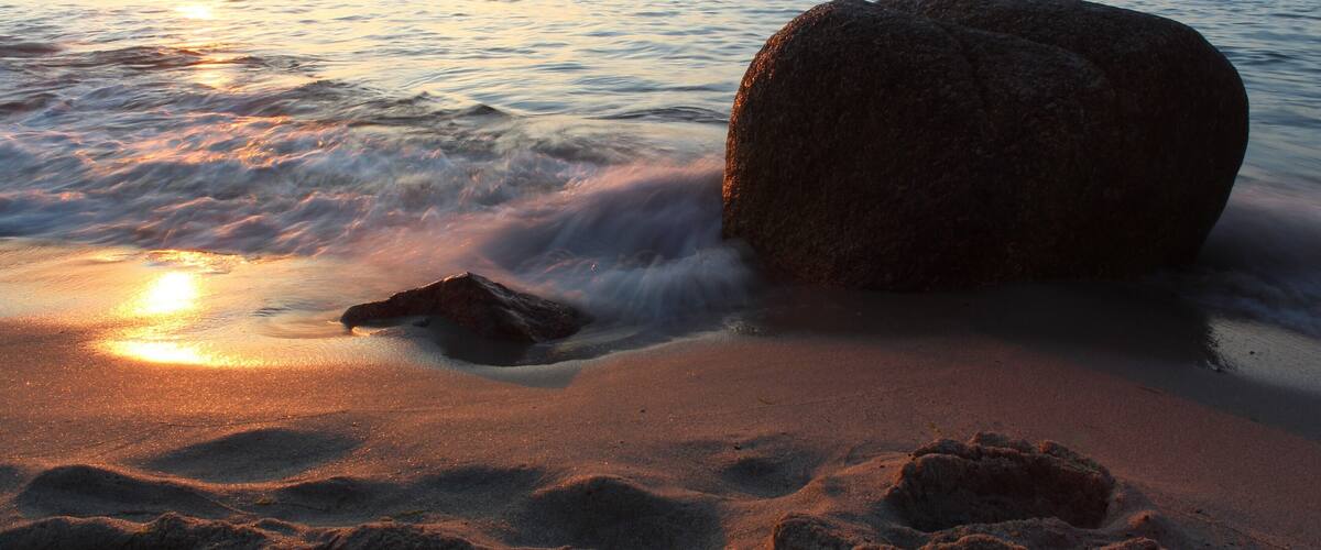 A beach with a large rock, waves, and a sunset.