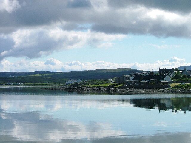 A coastal village with houses by the water, a calm sea, and hills in the background.
