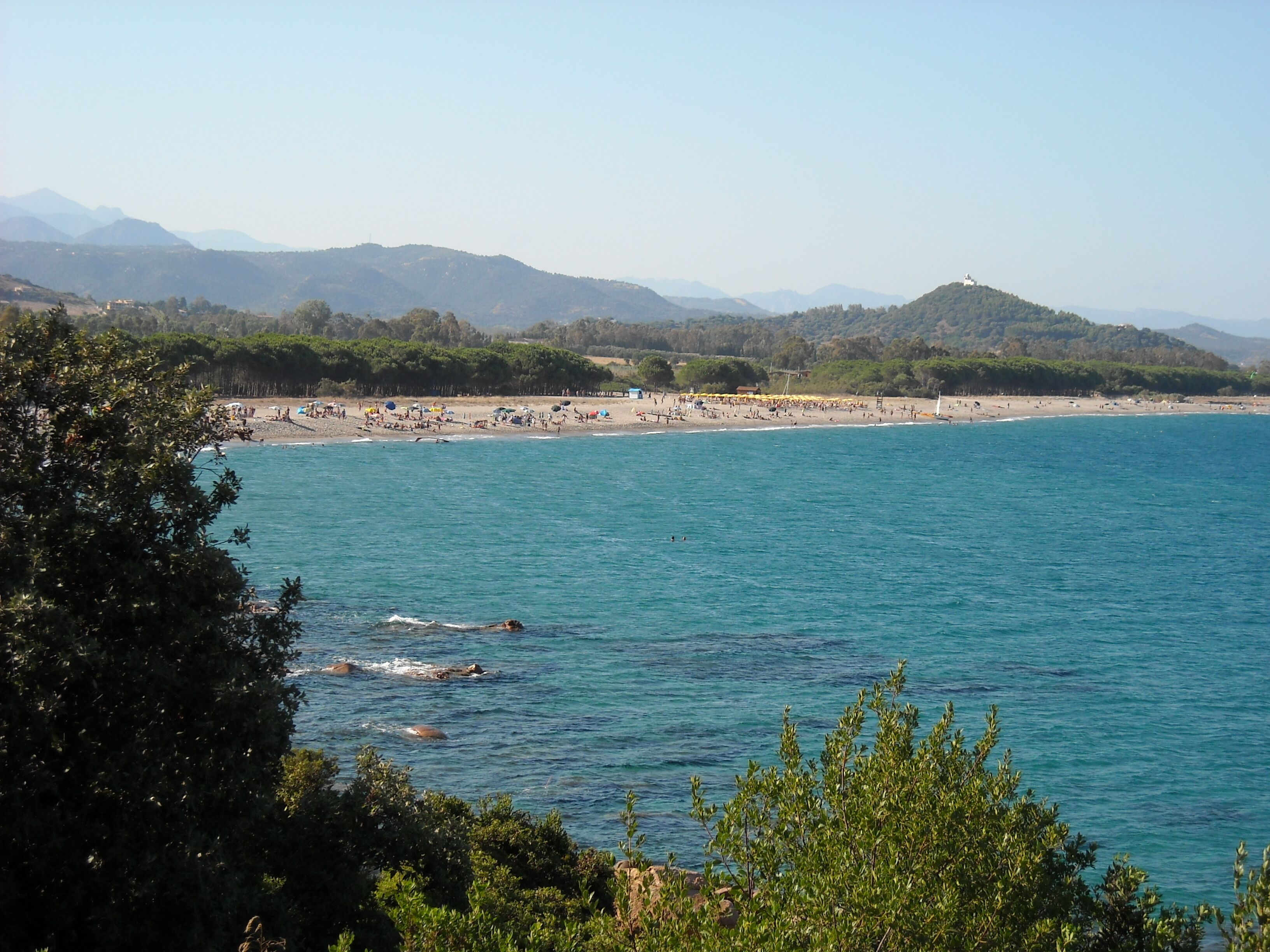 A coastal beach with clear blue waters, a sandy shore, and distant mountains.