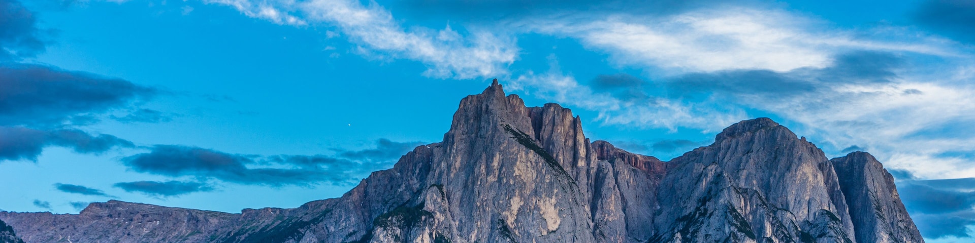 A mountainous landscape with a prominent rocky peak, a small village with traditional houses, and a clear blue sky with scattered clouds.