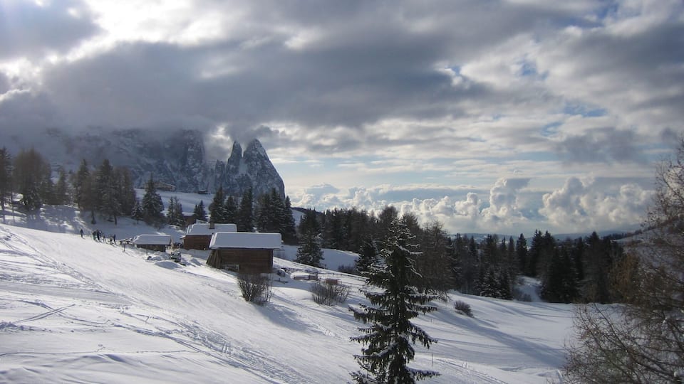A snowy mountain landscape with a cabin, pine trees, and a prominent peak in the background.