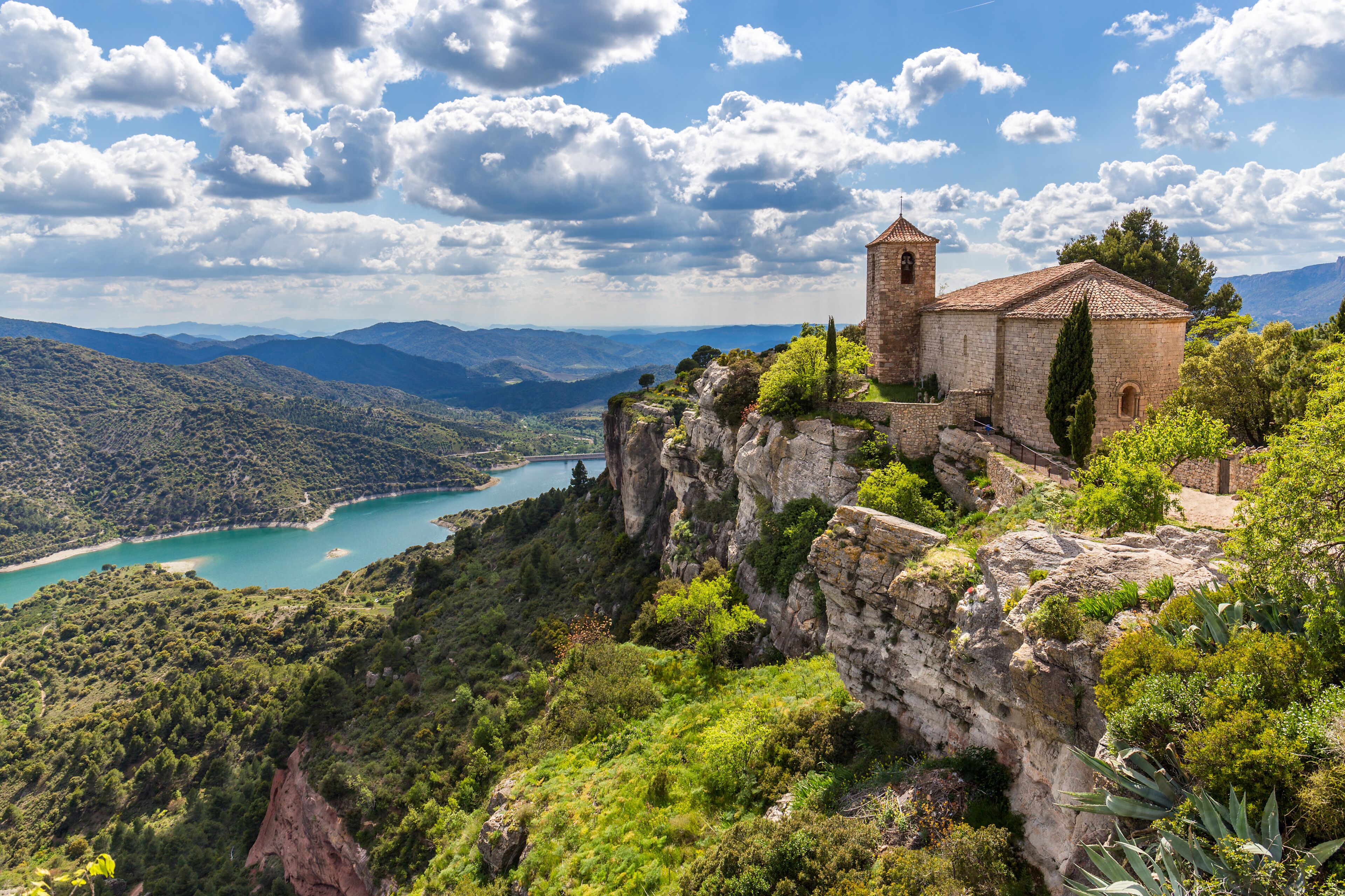 A historic stone church on a cliff overlooking a turquoise bay and lush green hills.