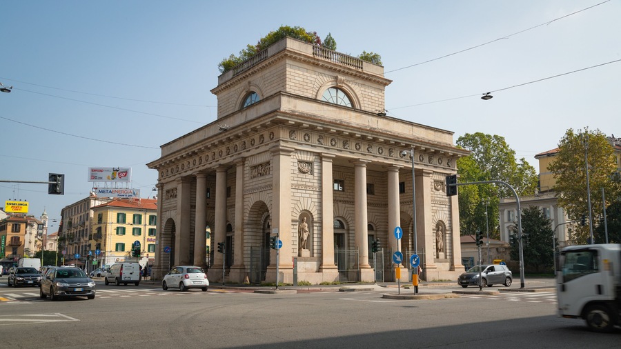 Un edificio storico dall'architettura classica, con colonne e statue, situato in un vivace incrocio cittadino.