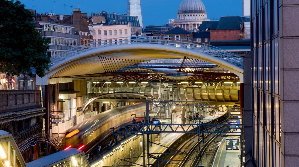 A modern train station with a curved roof, a moving train, and the London landmarks St. Paul's Cathedral and the Shard in the background.