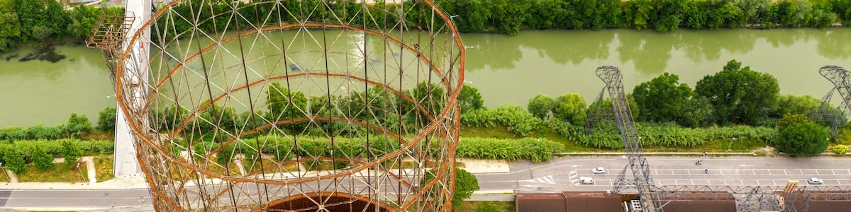 A large, circular metal structure with scaffolding, surrounded by greenery and a river.