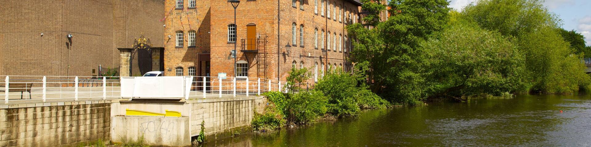 A historic brick building with a tower, a river, and lush greenery.
