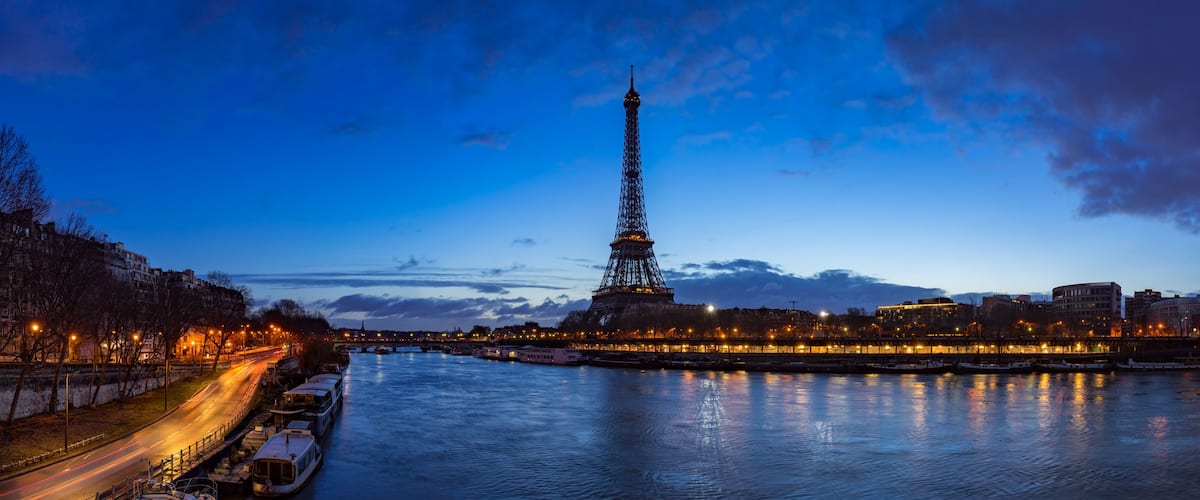 The Eiffel Tower at dusk with reflections on the Seine River, illuminated buildings, and a bridge in the background.