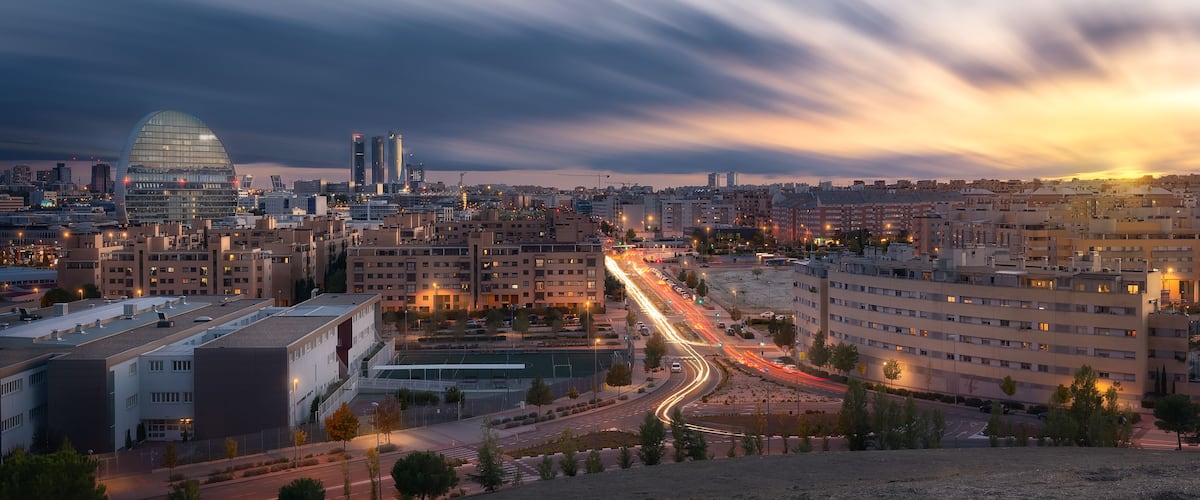 Un paisaje urbano al atardecer con un edificio curvo destacado, un campo deportivo y una calle concurrida con estelas de luz.