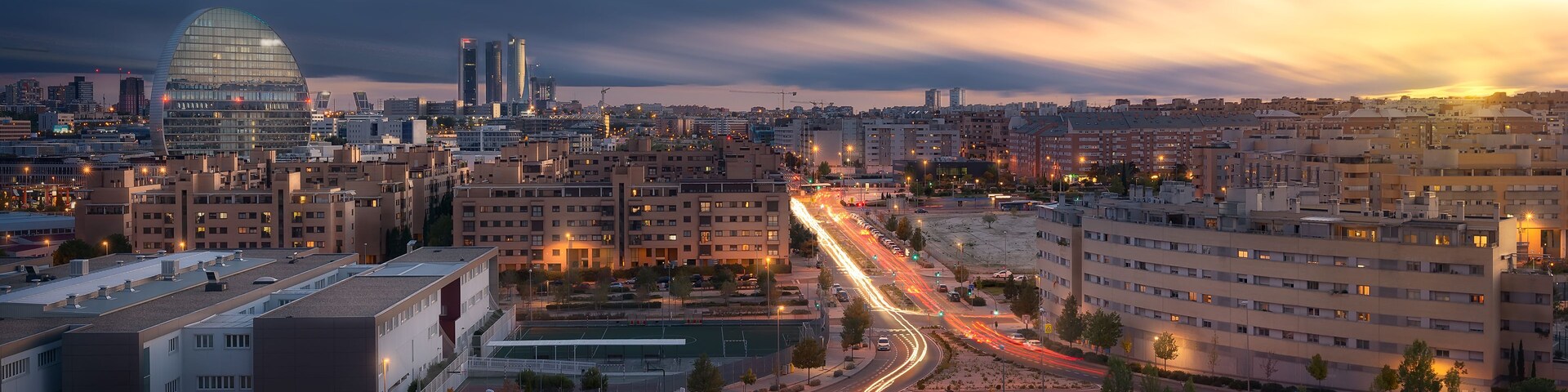 Un paisaje urbano al atardecer con un edificio curvo destacado, un campo deportivo y una calle concurrida con estelas de luz.