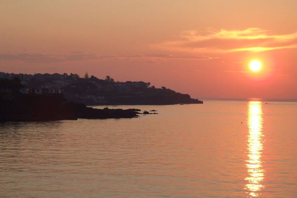 Een zonsondergang boven een rustige zee, met een silhouet van een kustplaatsje en rotsformaties.