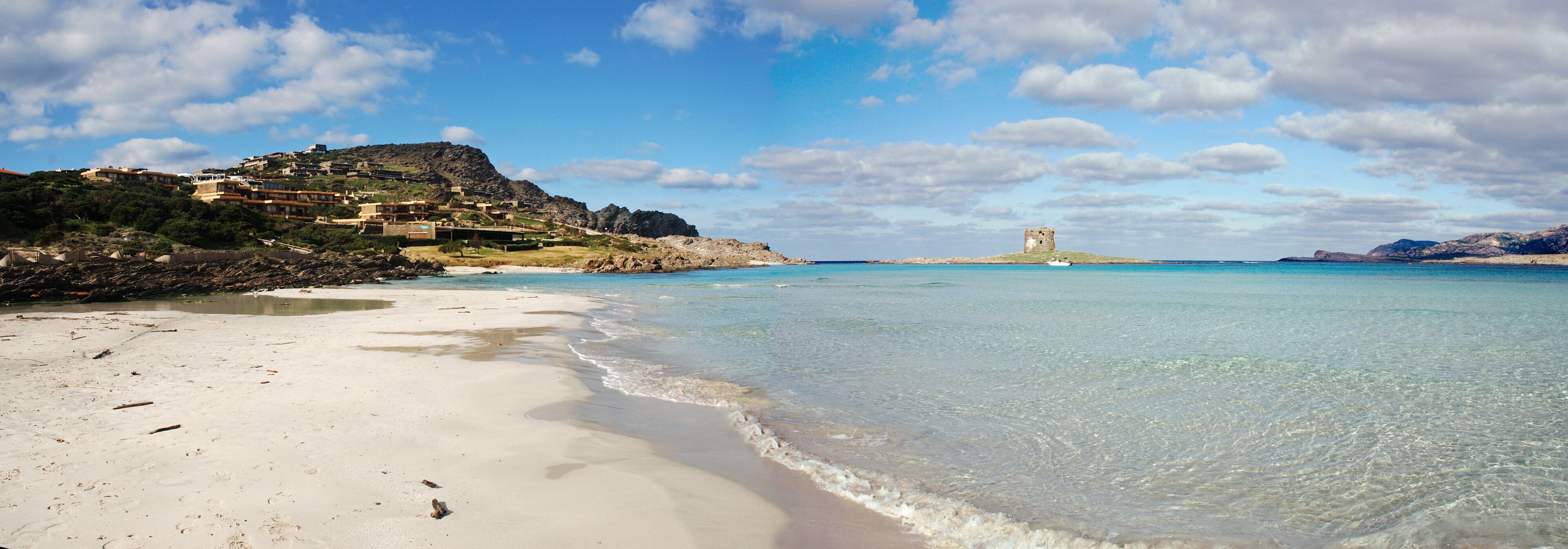 A beach with clear water, a castle on a hill, and buildings on the hillside.