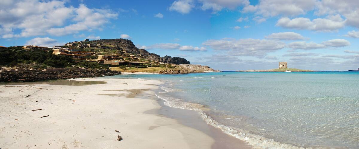 A beach with clear water, a castle on a hill, and buildings on the hillside.
