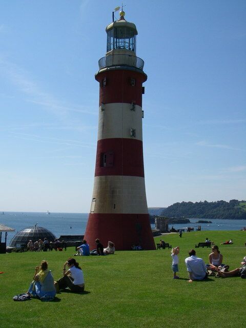 Un faro rojo y blanco con cielo despejado y una zona verde con gente.