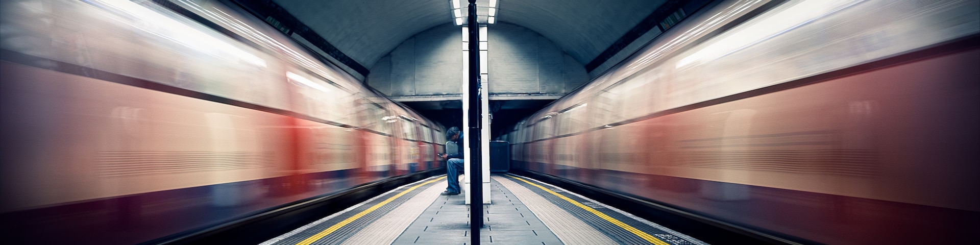 Estação de metro com trens em movimento oposto, um pilar central e piso tátil amarelo na plataforma.