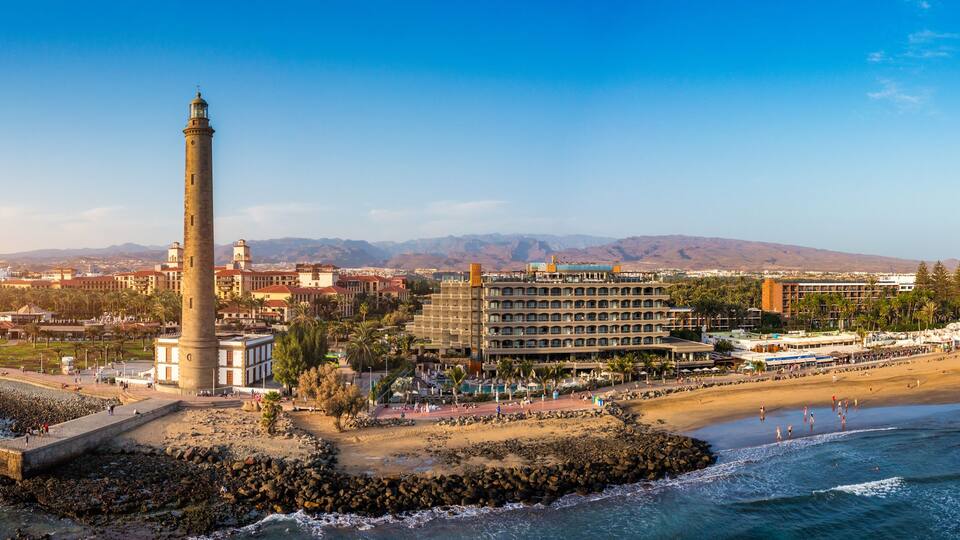 Un paisaje costero con un faro destacado, una playa y montañas al fondo.