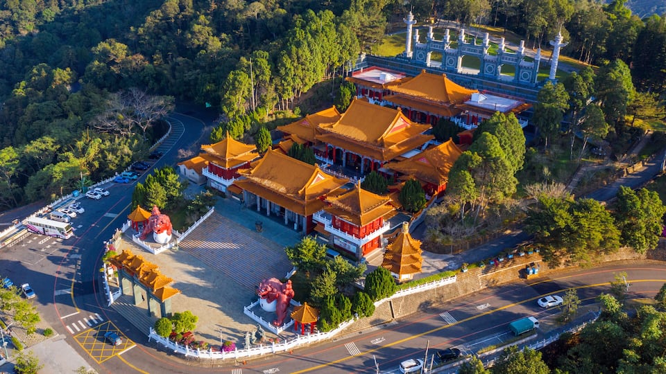 An aerial view of a traditional Chinese temple complex with multiple buildings, a large red lion statue, and a surrounding road.
