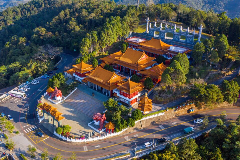 An aerial view of a traditional Chinese temple complex with multiple buildings, a large red lion statue, and a surrounding road.