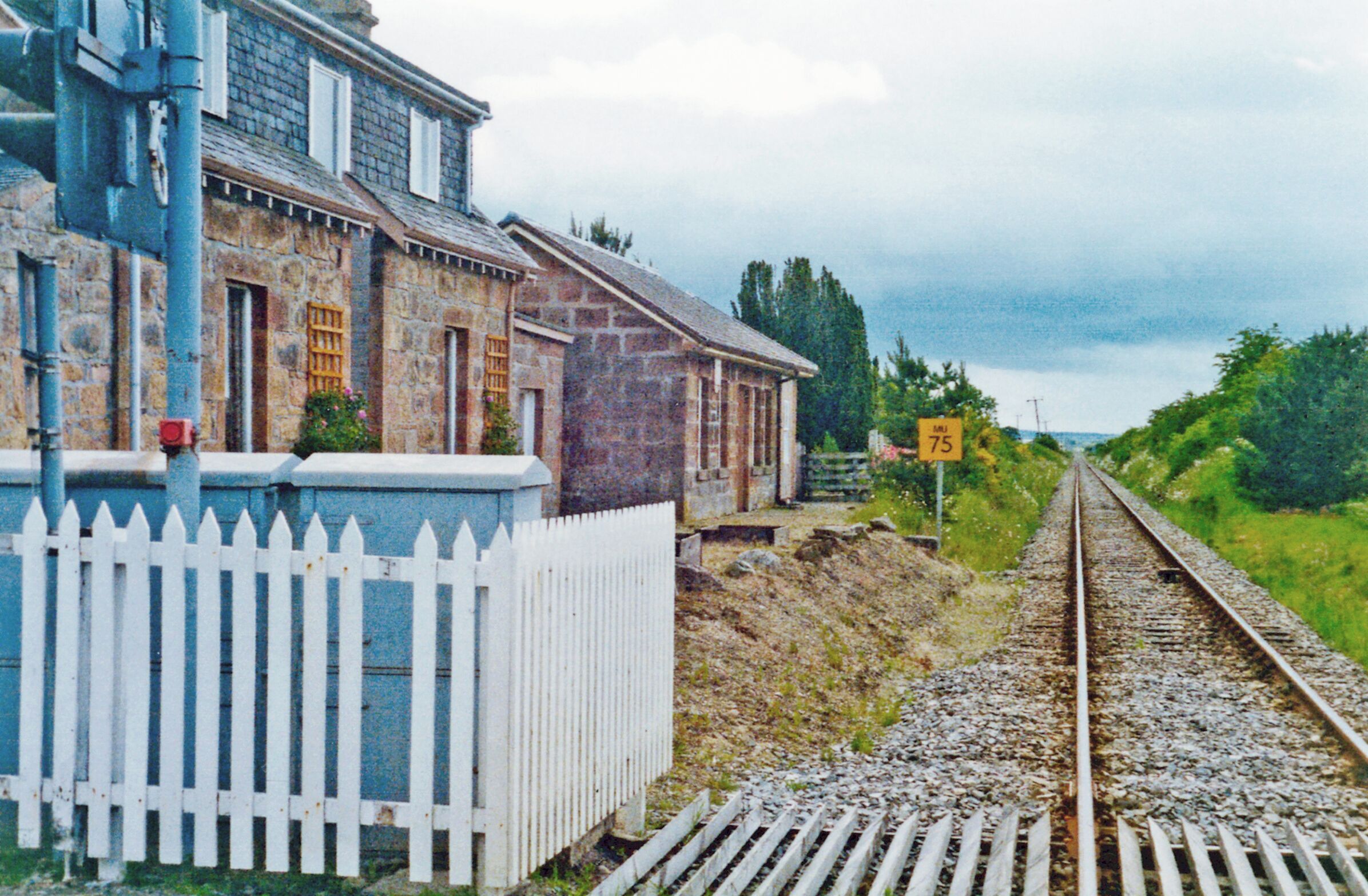 A stone building with a white picket fence, a railway track, and a speed limit sign of 75.