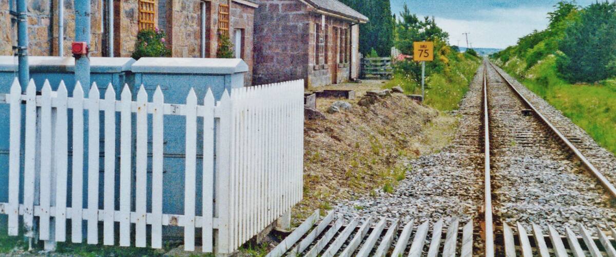 A stone building with a white picket fence, a railway track, and a speed limit sign of 75.