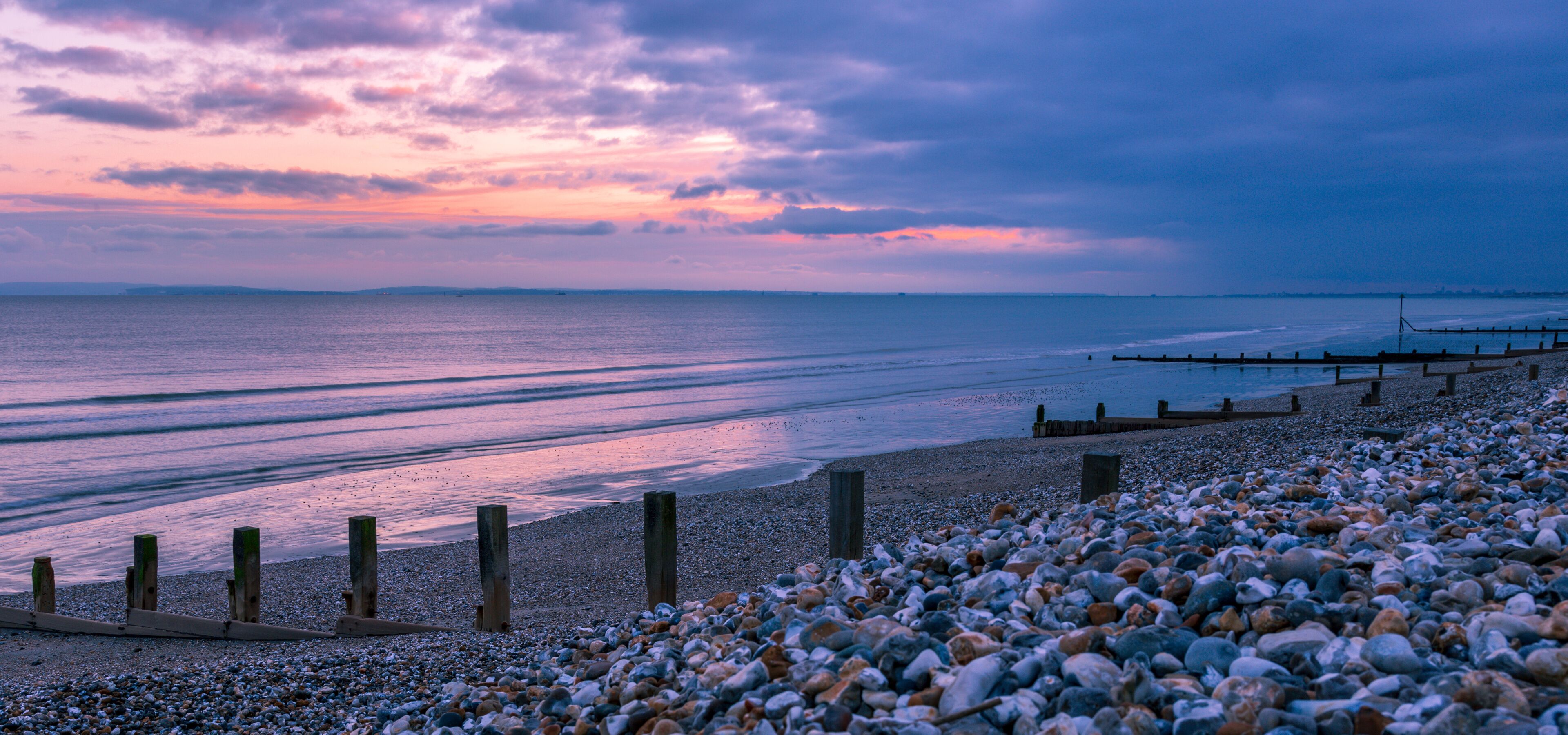 A pebbled beach with wooden posts and a wooden jetty extending into the water.