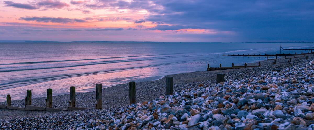 A pebbled beach with wooden posts and a wooden jetty extending into the water.
