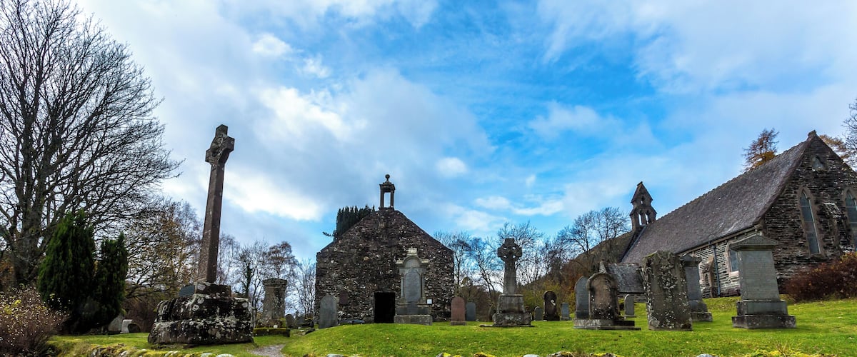 Eine historische Steinkirche mit hohem Glockenturm, umgeben von einem Friedhof mit Grabsteinen und einer Steinmauer.