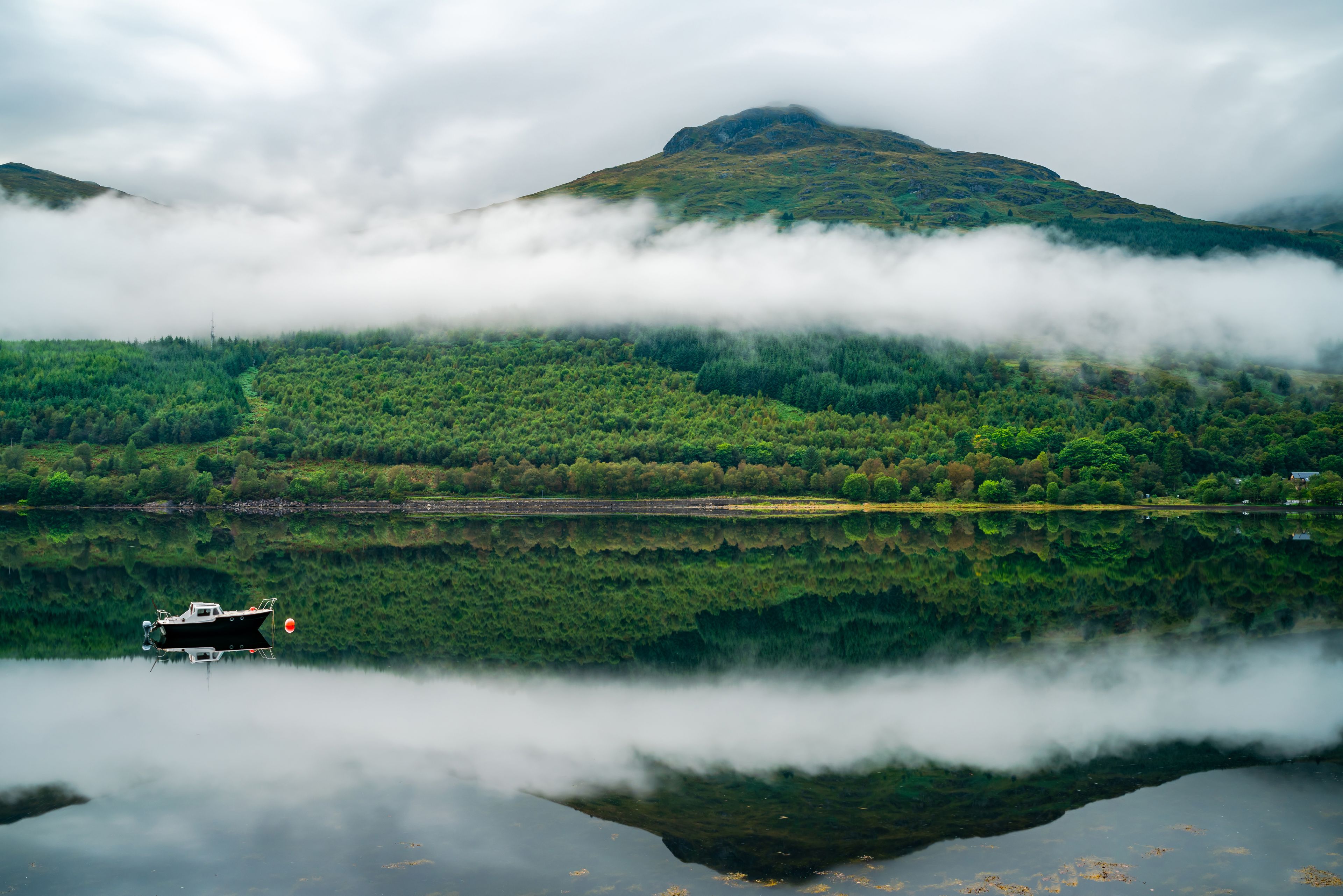 Un lago tranquillo con una barca, una fitta foresta e una montagna parzialmente nascosta dalle nuvole.