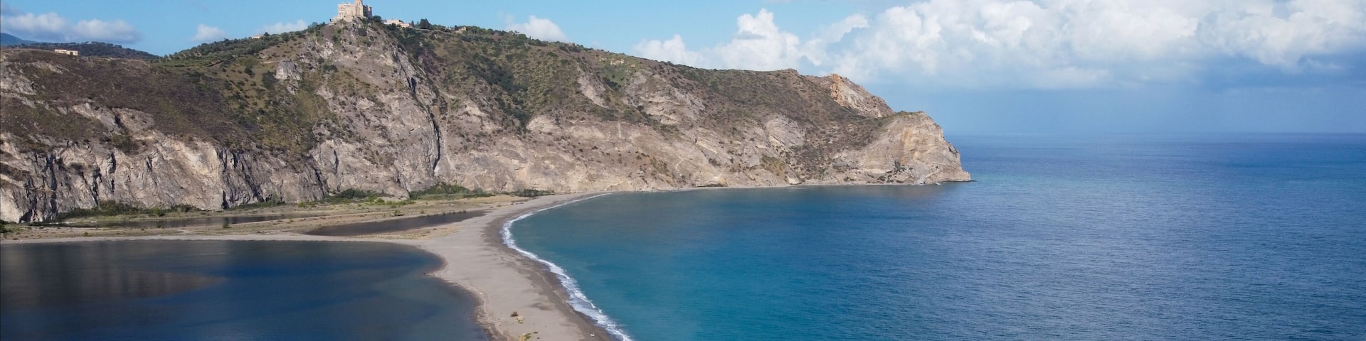 A coastal landscape with a sandy beach, a clear blue sea, and a rocky hill with a structure on top.