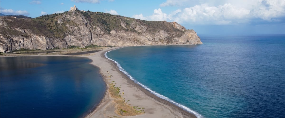 A coastal landscape with a sandy beach, a clear blue sea, and a rocky hill with a structure on top.