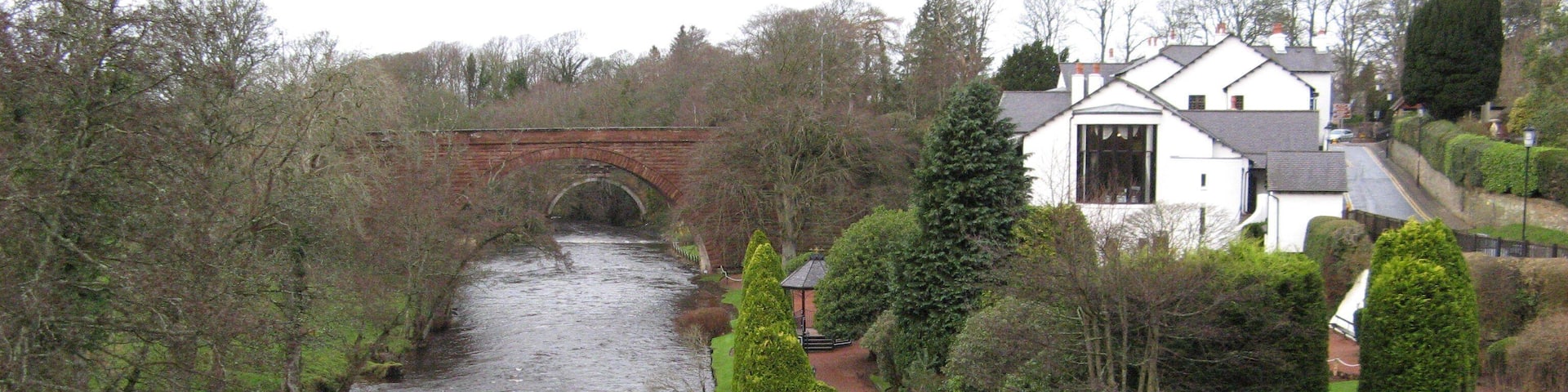 A river flowing under a bridge, surrounded by neatly trimmed hedges and a white building with a sloped roof.