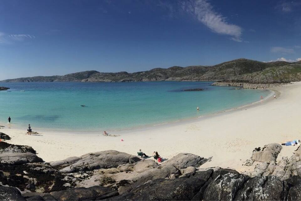 Uma praia com pedras em primeiro plano, águas turquesas translúcidas e uma faixa de areia.