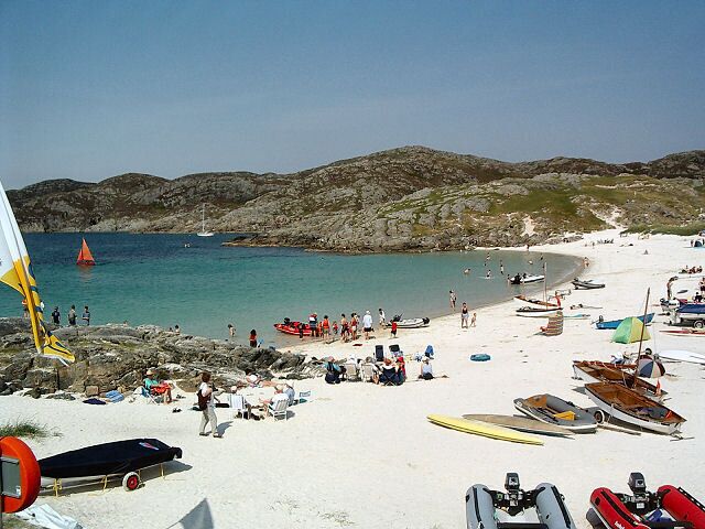 A beach with boats, people, and a sailboat in the water.