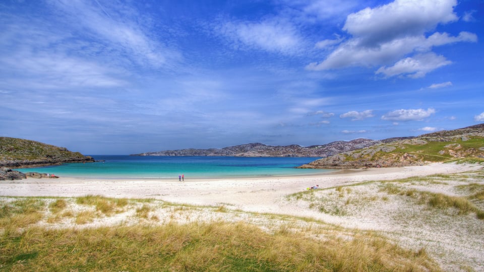 A coastal landscape with a sandy beach, grassy dunes, and a clear blue sea under a partly cloudy sky.