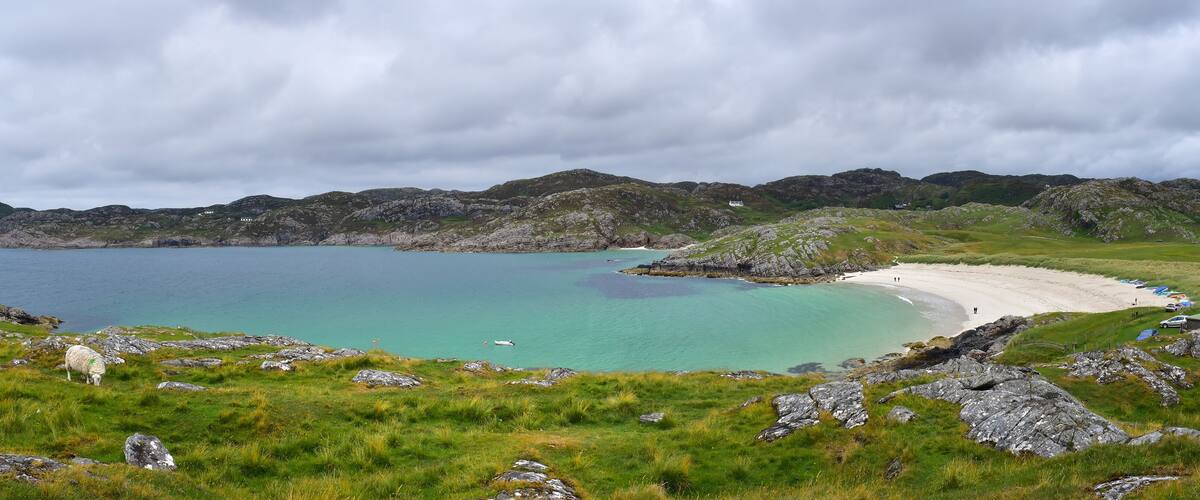 A coastal landscape with a sandy beach, turquoise waters, rocky hills, and a grassy foreground.